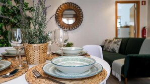 Un coin repas avec une table dressée pour deux personnes, agrémenté de sets de table en fibres tressées, de bols en verre transparent et de verres à vin. Un miroir et un canapé vert sont visibles à l’arrière-plan.