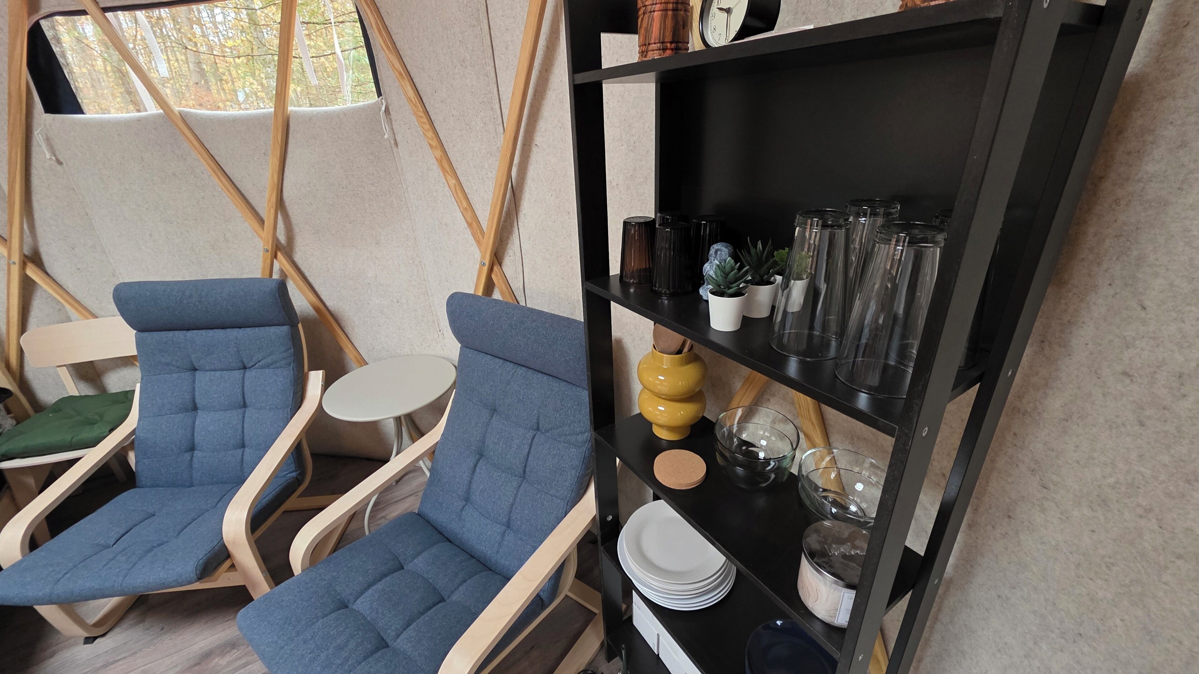 Ein Zimmer mit zwei blauen Sitzgelegenheiten, einem kleinen runden Tisch, einer schwarzen Regalanlage mit Gläsern und einem Fenster mit Blick auf Bäume.