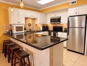 A kitchen with white cabinets, a stainless steel refrigerator, a microwave, and a large island with stools. - Saida III Condos - S3706 (South Padre Island)