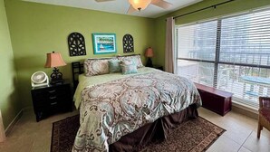 A bedroom with a bed, a ceiling fan, a nightstand, a lamp, a window with blinds, and a view of the outside. - Saida III Condos - S3706 (South Padre Island)