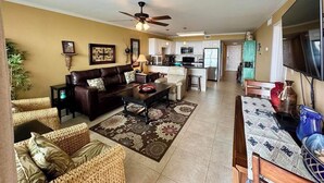 A spacious living room with a dark brown sofa, a coffee table, and a patterned rug. A ceiling fan is visible, and the room has a neutral color palette. - Saida III Condos - S3706 (South Padre Island)