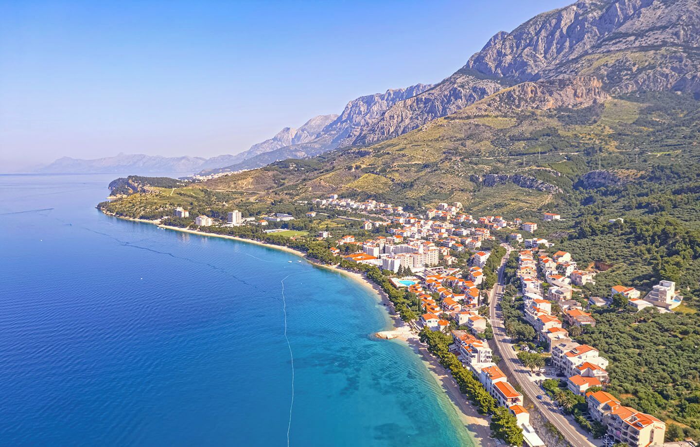 An aerial view of a coastal town with a clear blue sea, a beach, and mountainous terrain in the background.