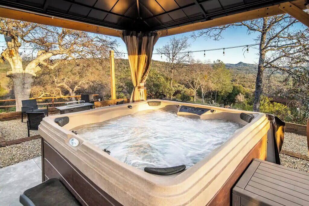 Un jacuzzi con terraza de madera y zona de estar al aire libre, rodeado de árboles y colinas.