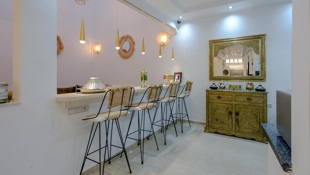 A dining area with a long table, bar stools, a decorative mirror, and a cabinet with tea sets. - Dar Mawati (Marrakech)