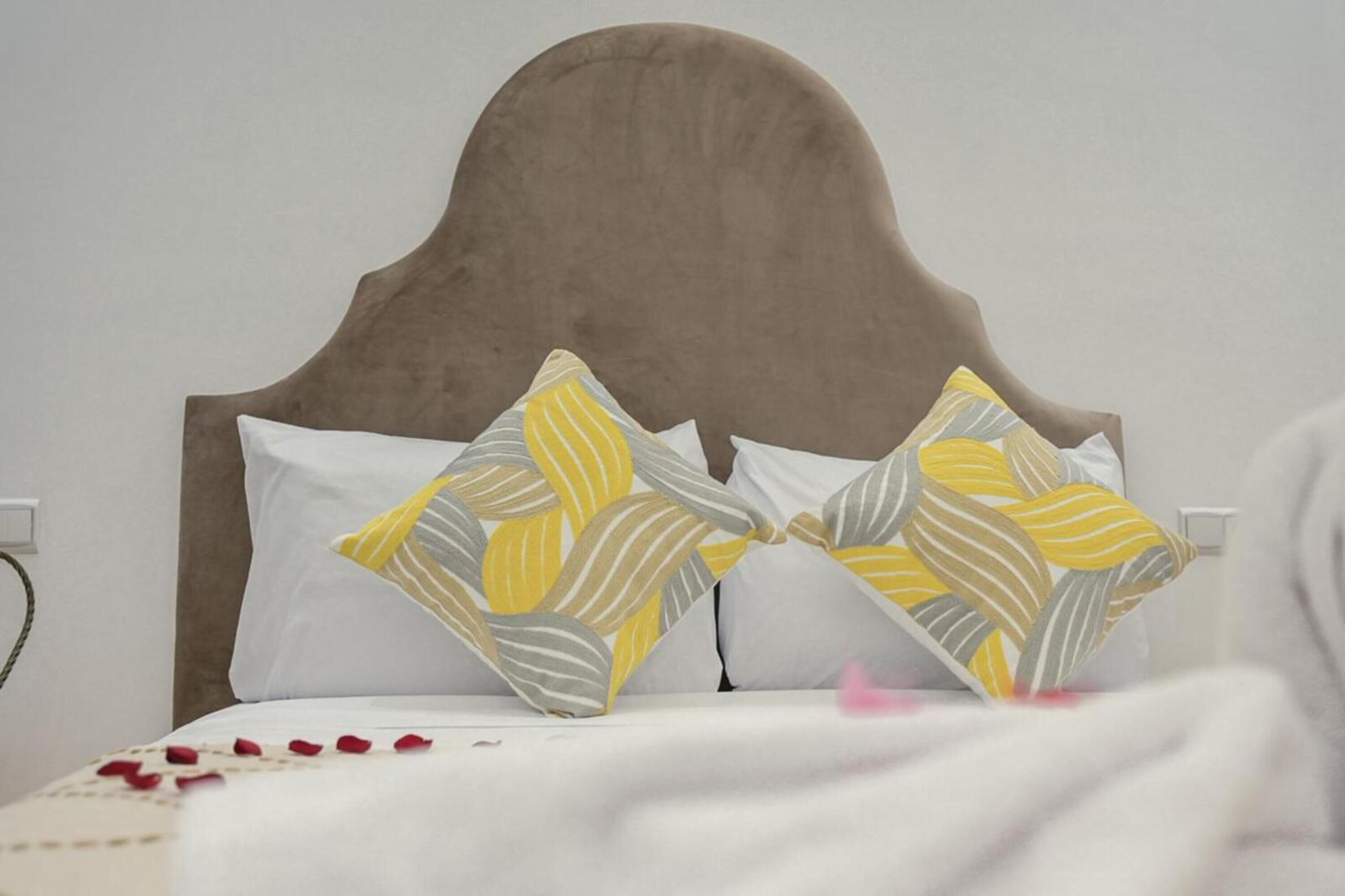 Un letto con una testiera marrone, cuscini bianchi e una coperta con petali di rosa rossa.