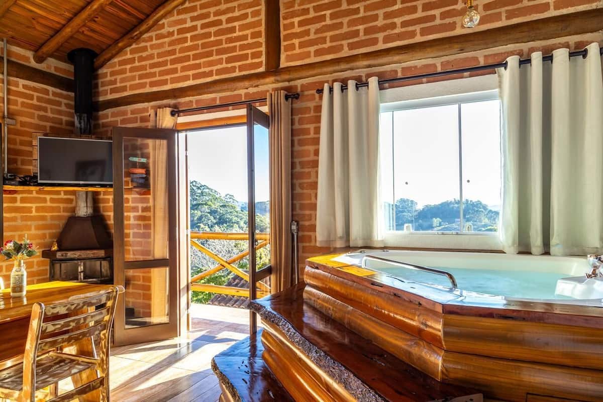 A rustic cabin interior with a wooden table, chairs, a fireplace, a large window with curtains, and a hot tub.