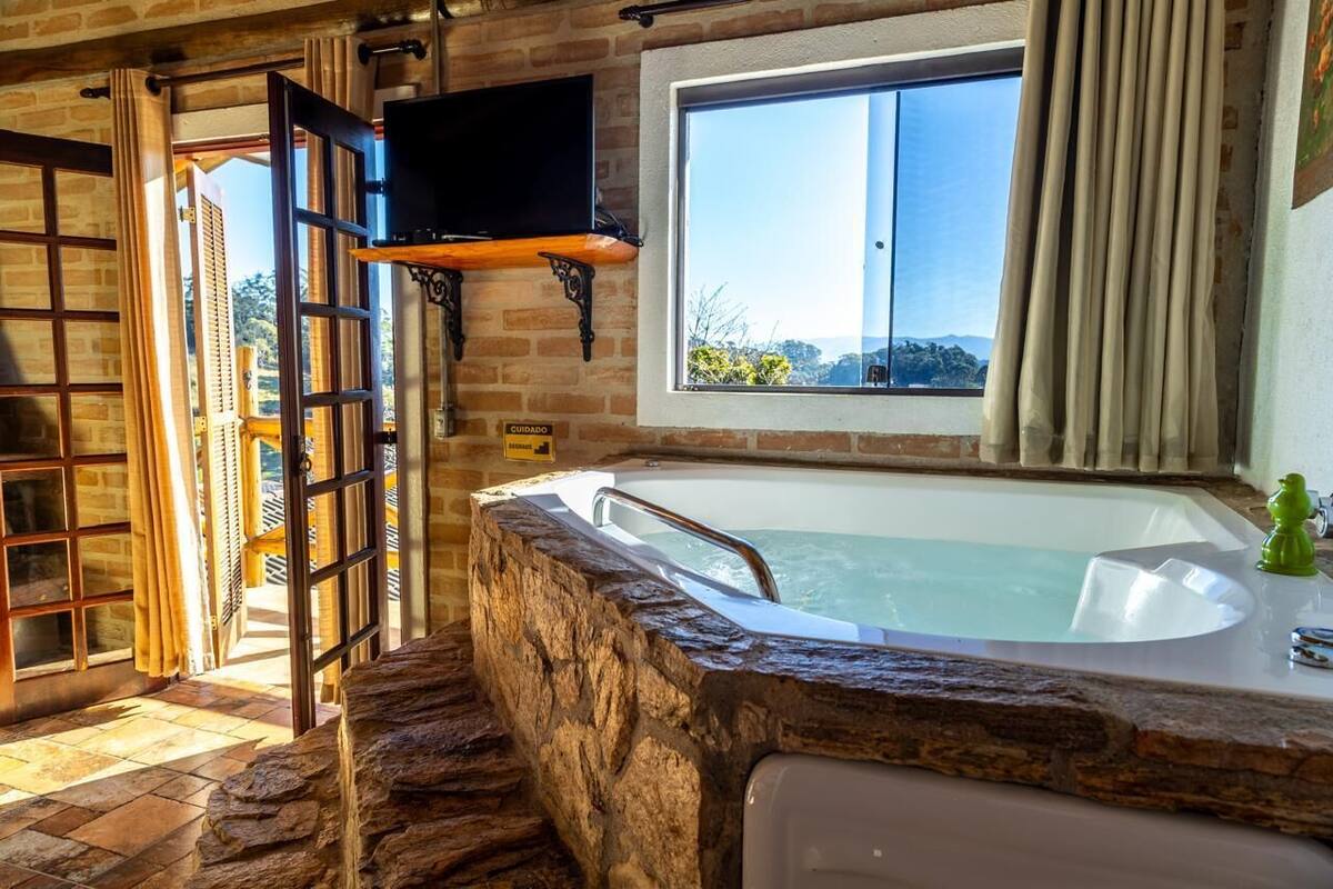 A bathroom with a stone wall, a bathtub, a window, and a television mounted above the tub.