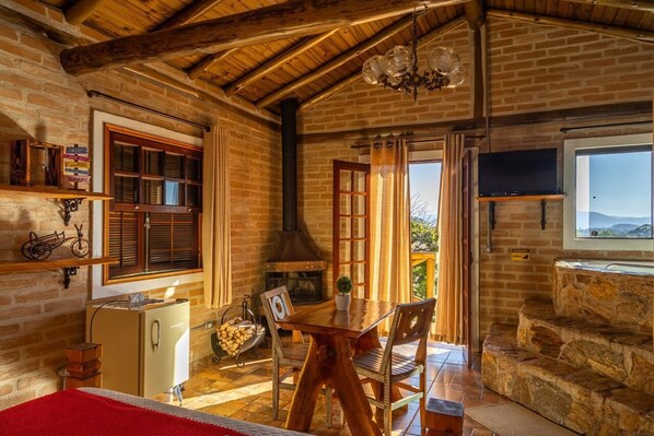 Een rustieke kamer met een bakstenen muur, houten balken in het plafond, een open haard, een eettafel met stoelen en een aan de muur bevestigde televisie.