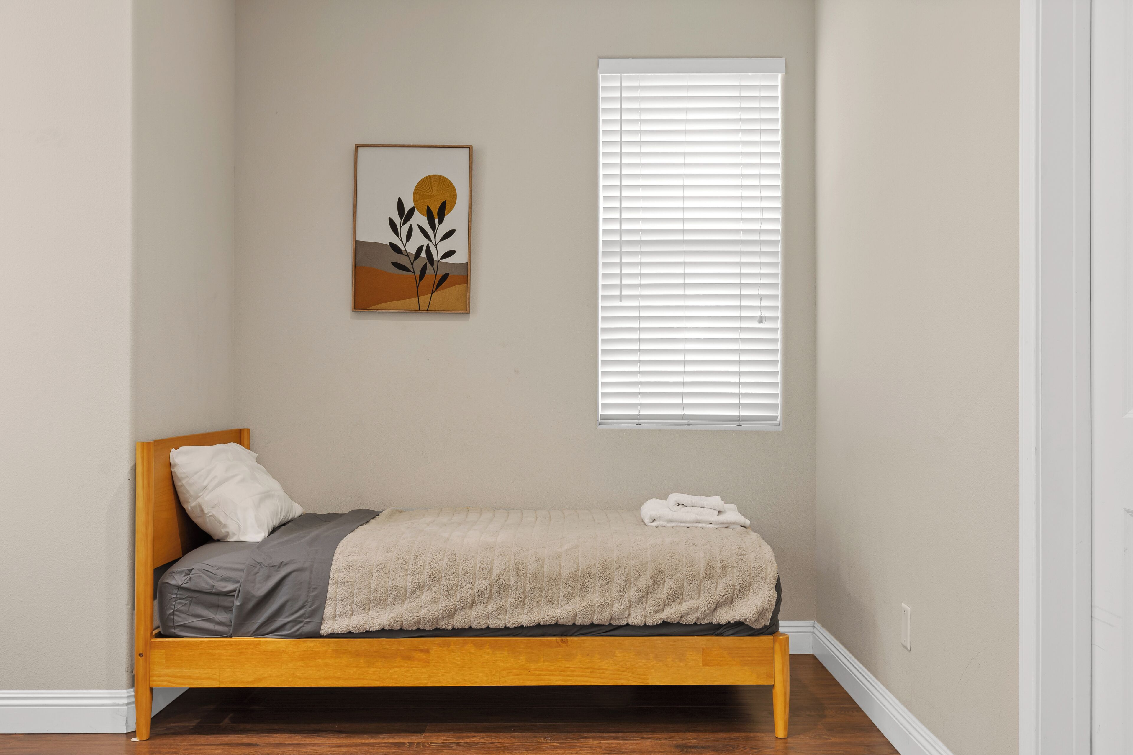 Una cama individual con estructura de madera, colchón gris y manta color beige. Hay una toalla blanca sobre la cama. Un cuadro enmarcado cuelga en la pared sobre la cama.