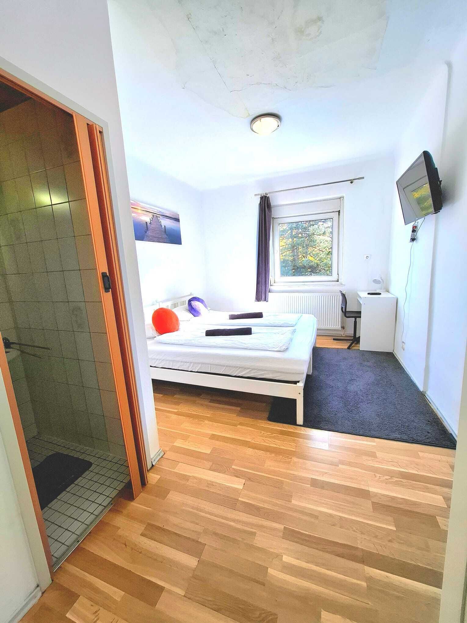 A small, well-lit room with a bed, a window, a TV, and a radiator.