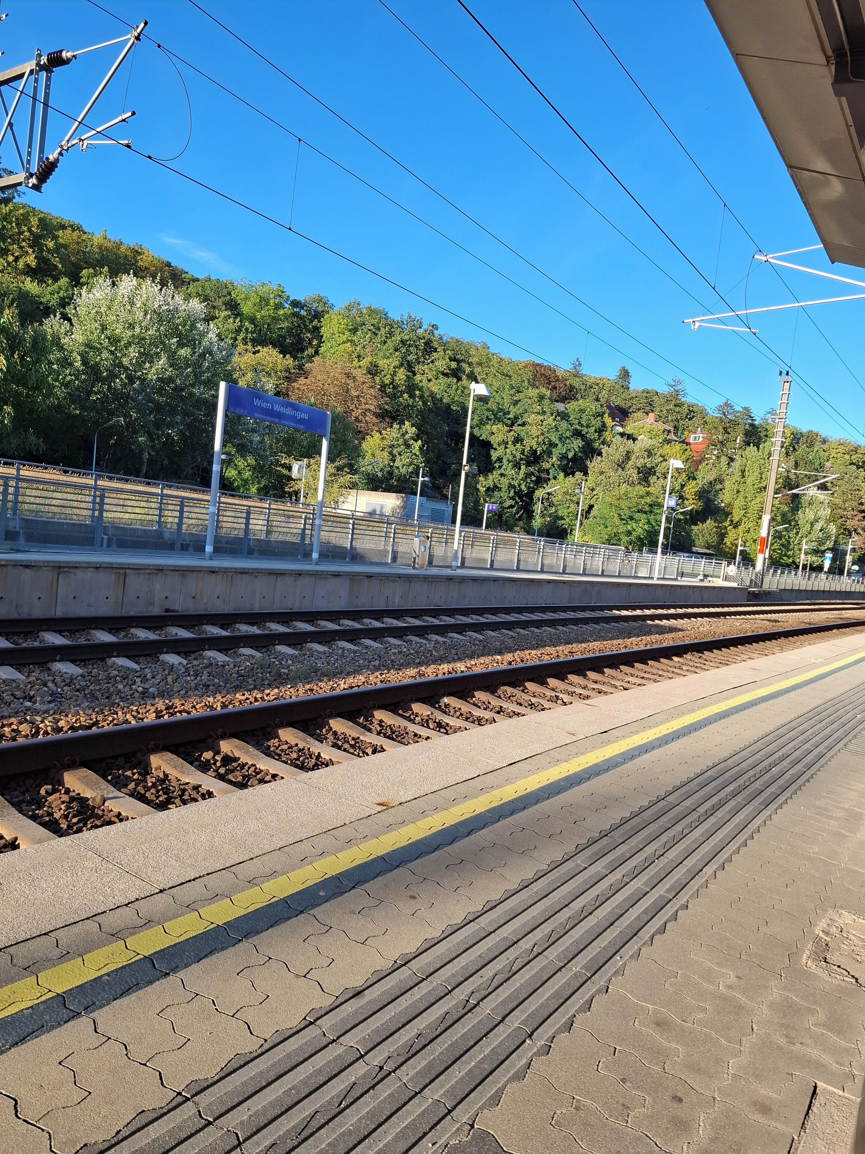Een treinperron met een heldere lucht, een bordje met Wien Weidlingau, en een glooiend landschap op de achtergrond.