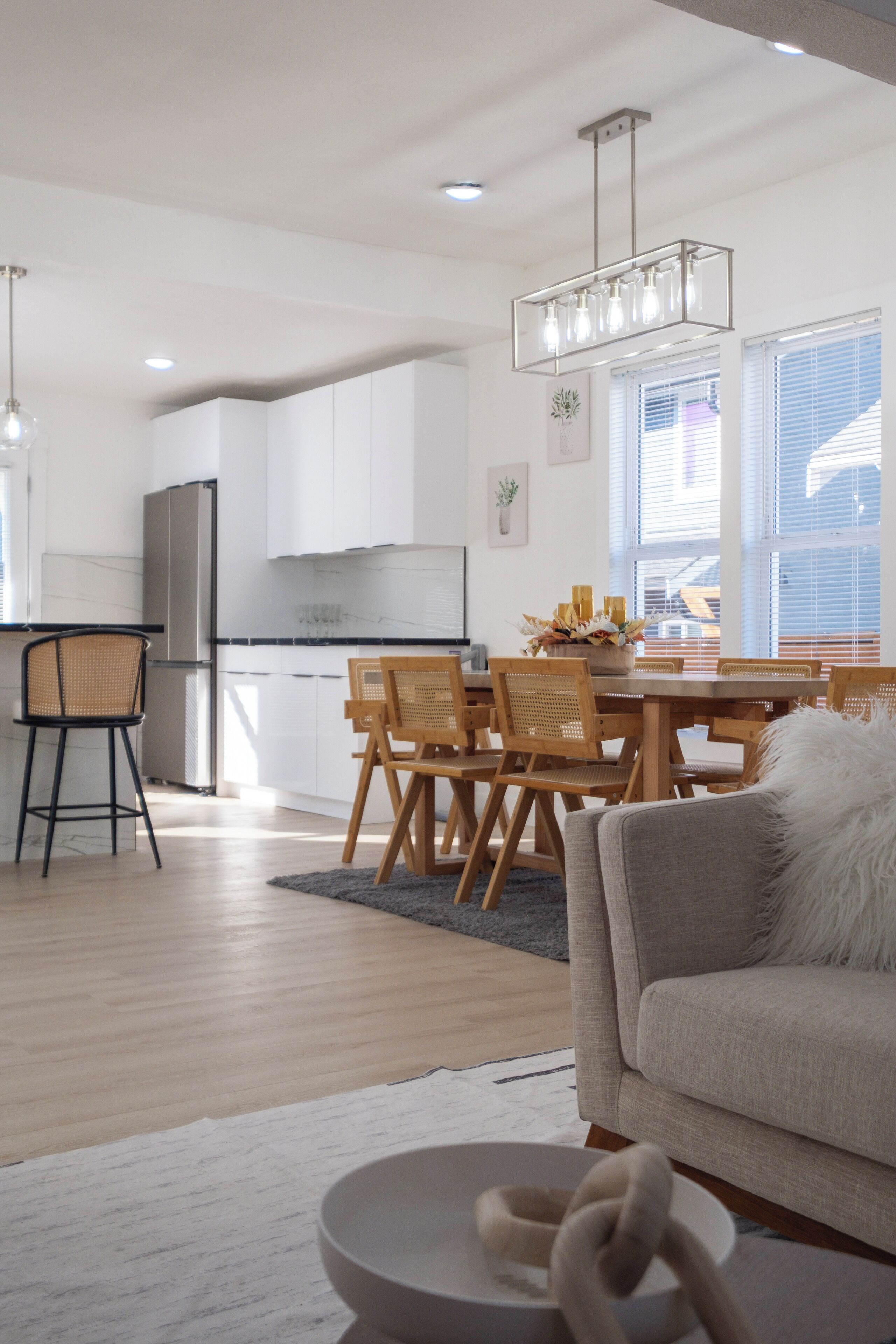 A modern kitchen with white cabinets, a dining area with wooden chairs, and a grey sofa in the living room.