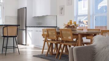 A modern kitchen with white cabinets, a dining area with wooden chairs, and a grey sofa in the living room.