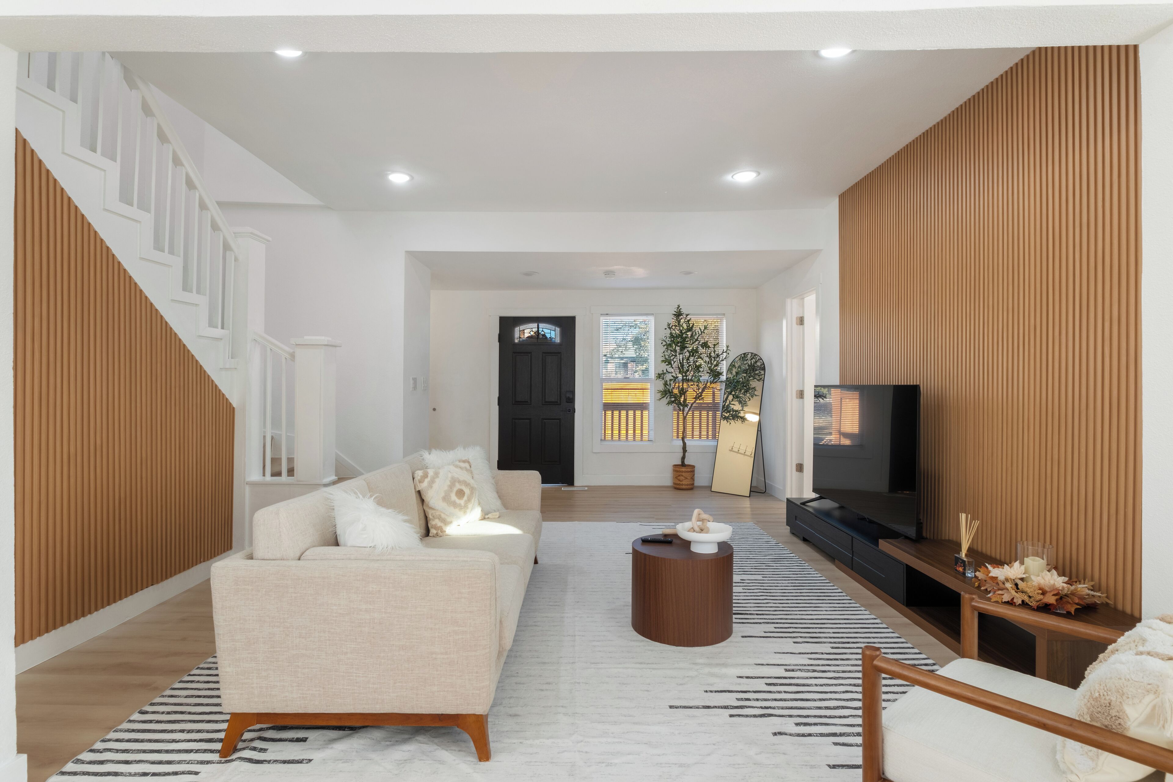 A modern living room with a sofa, coffee table, and TV. A staircase is visible on the left, and a wooden panel wall is on the right.