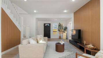 A modern living room with a sofa, coffee table, and TV. A staircase is visible on the left, and a wooden panel wall is on the right.
