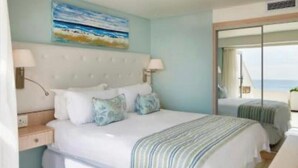 A bedroom with a bed, bedside tables, a painting, and a view of the sea.