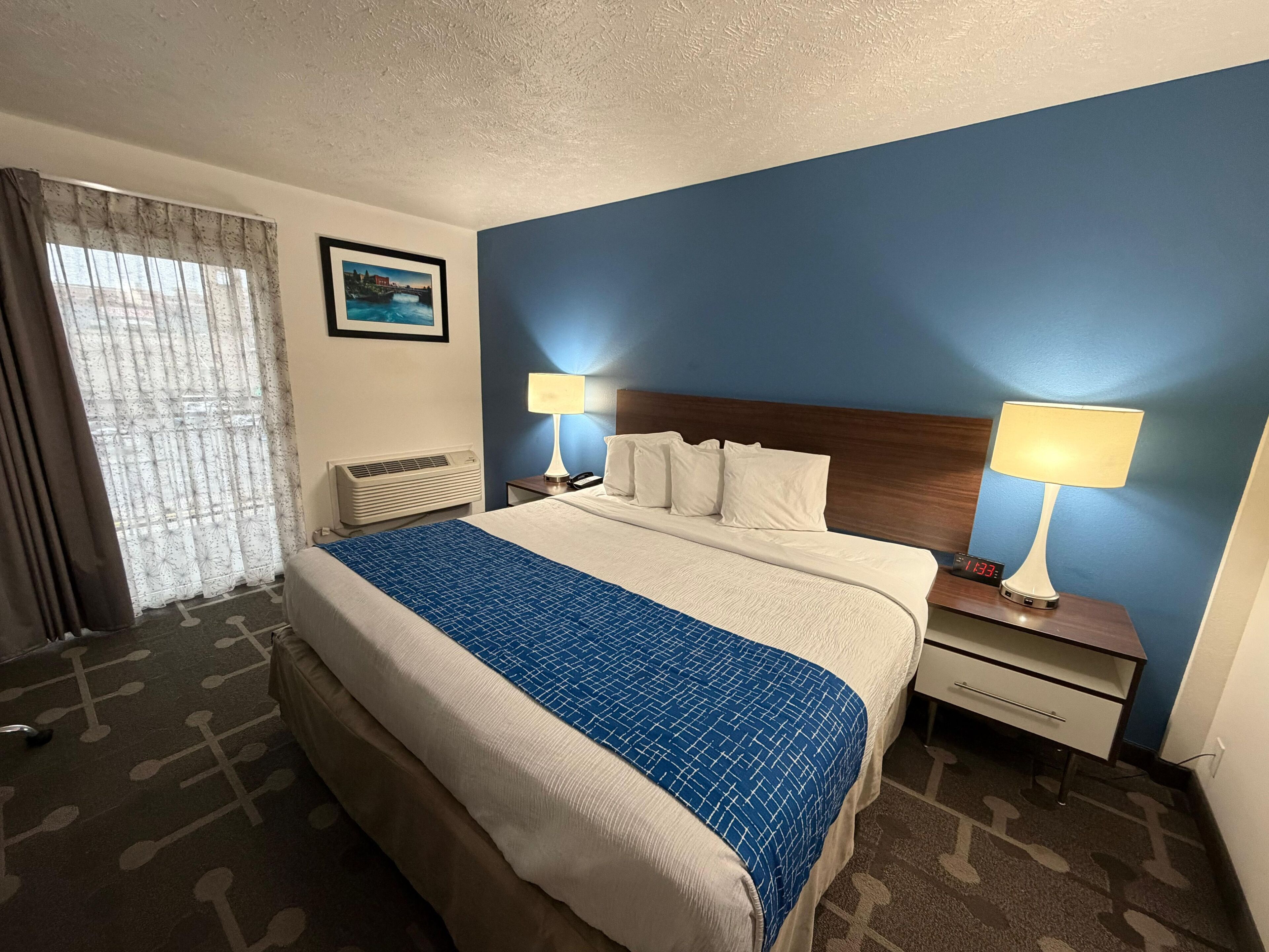 Habitación de hotel con cama, dos mesitas de noche con lámparas, un calentador, ventana con cortinas y un cuadro en la pared.