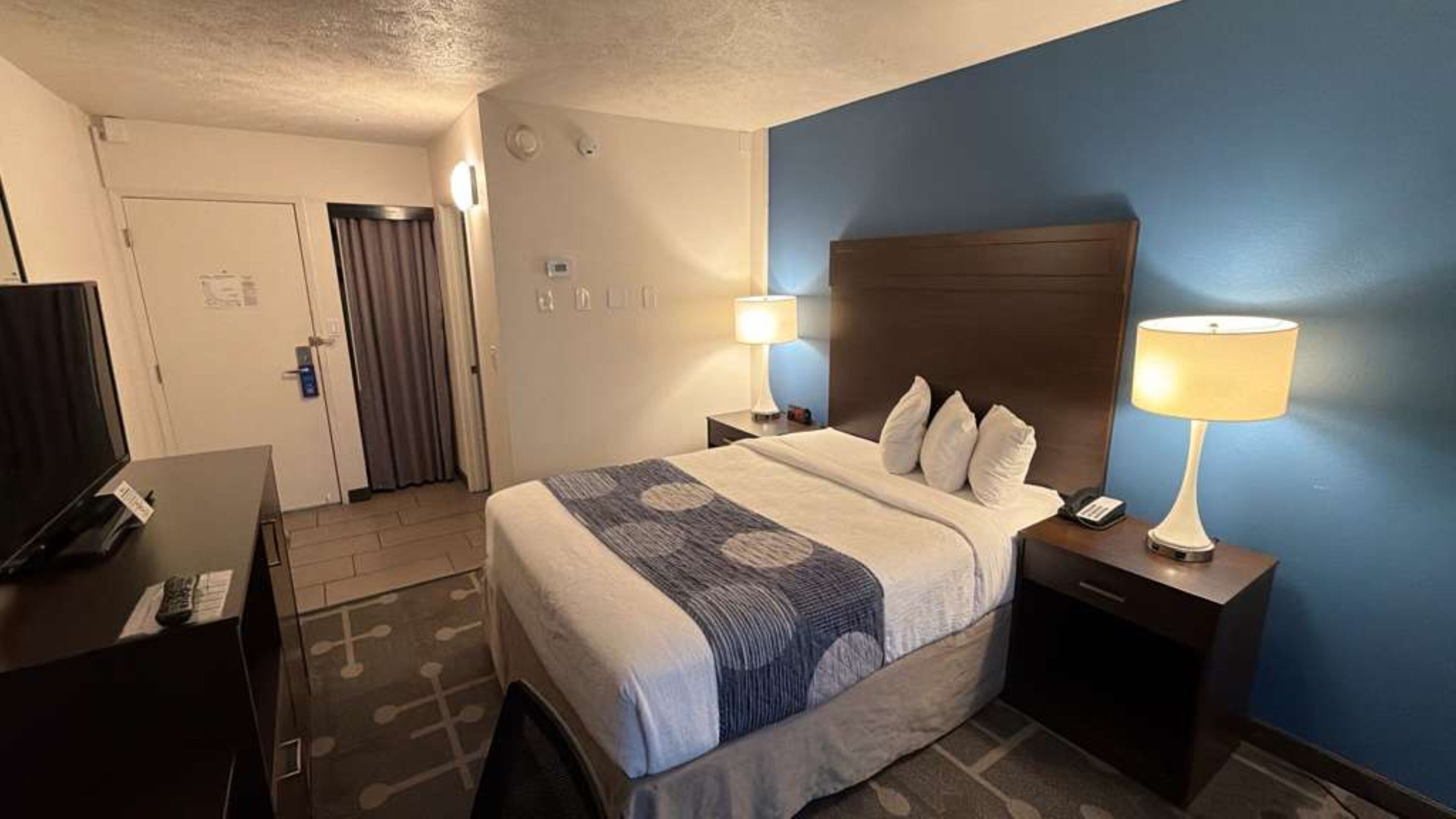 A hotel room with a bed, two bedside lamps, a TV, and a door with curtains.