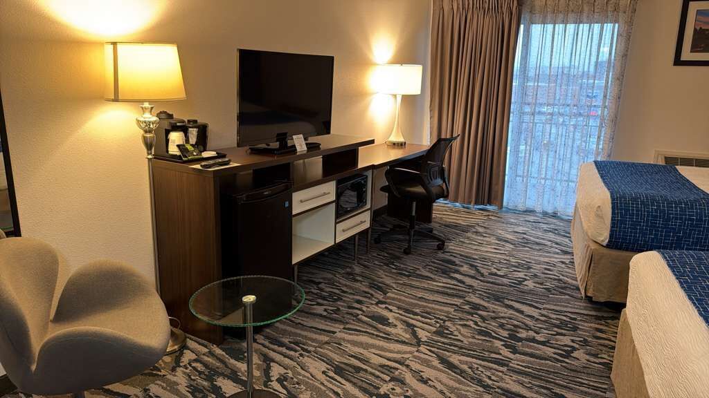 A hotel room with a bed, a desk with a TV, a chair, a glass table, and a view of the city through the window.