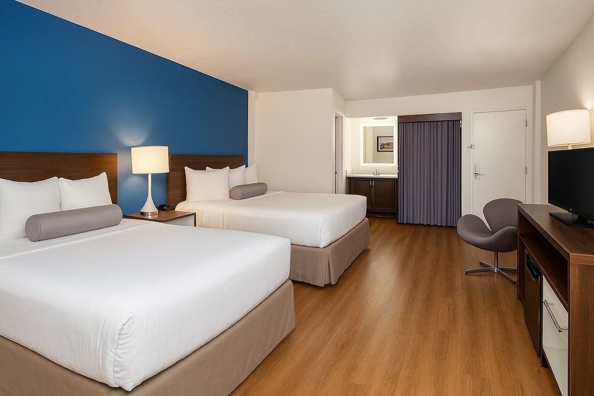 Habitación de hotel con dos camas, un escritorio con televisor, una silla y un baño visible a través de una puerta abierta.