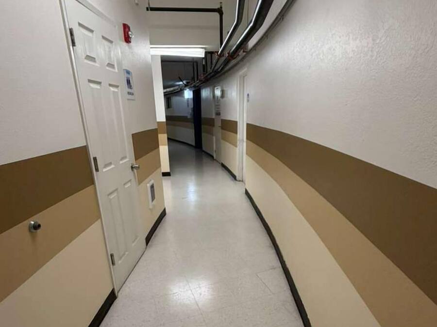 Um corredor com paredes bege e brancas, uma porta branca e um extintor de incêndio na parede.