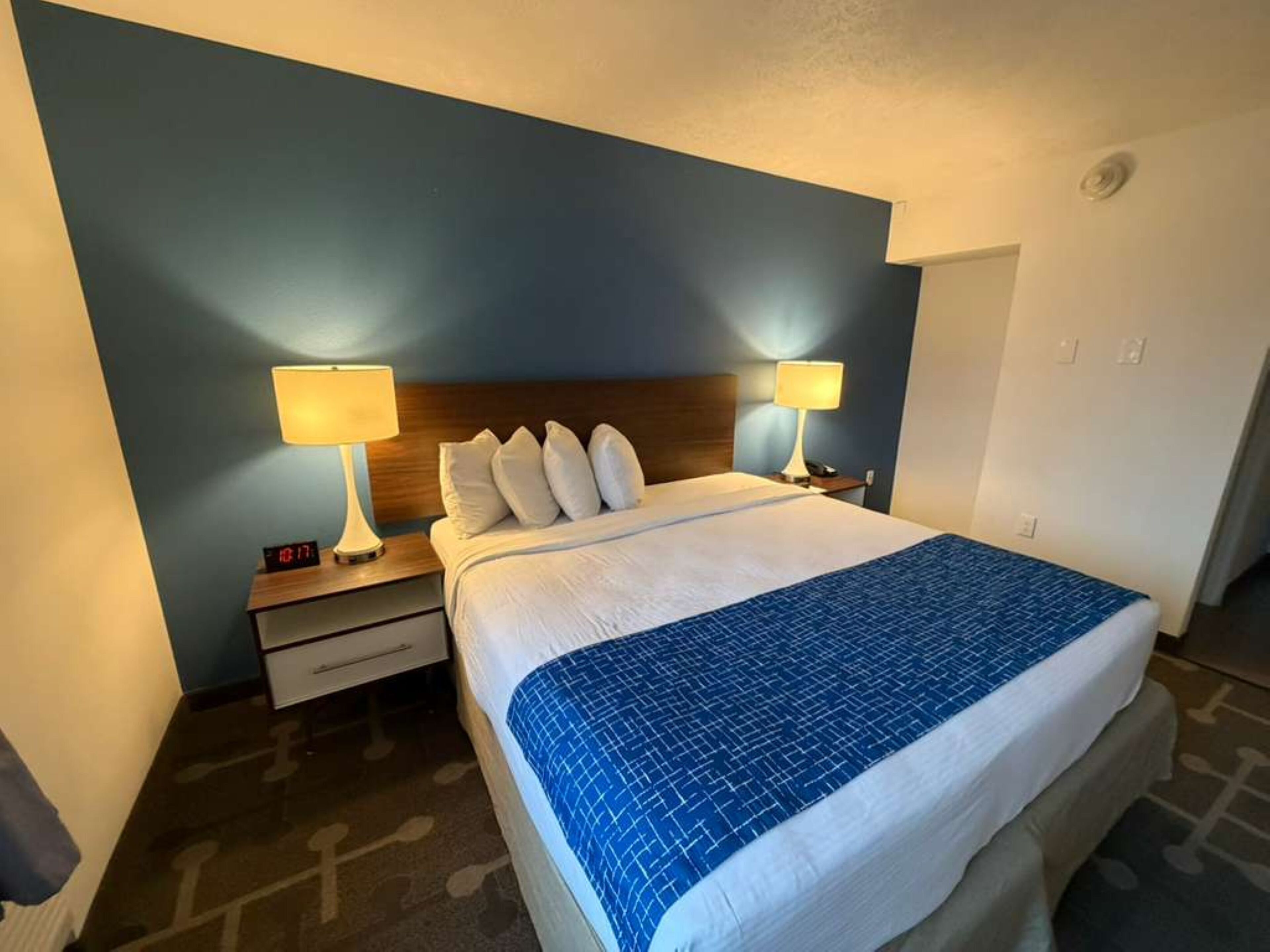 Habitación de hotel con cama, dos lámparas de noche, una mesita de noche y un reloj digital.