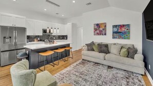 A modern kitchen with white cabinets, stainless steel appliances, and a central island with stools. There is a gray sofa and a patterned rug in the living area. - HTX NRG + Med Center (Houston)