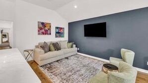 A modern living room with a grey accent wall, a flat-screen TV mounted above a sofa, and a patterned rug. - HTX NRG + Med Center (Houston)