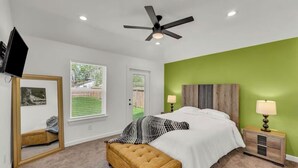 A bedroom with a bed, a ceiling fan, a nightstand with a lamp, a mirror, and a window with a view of the outdoors. - HTX NRG + Med Center (Houston)