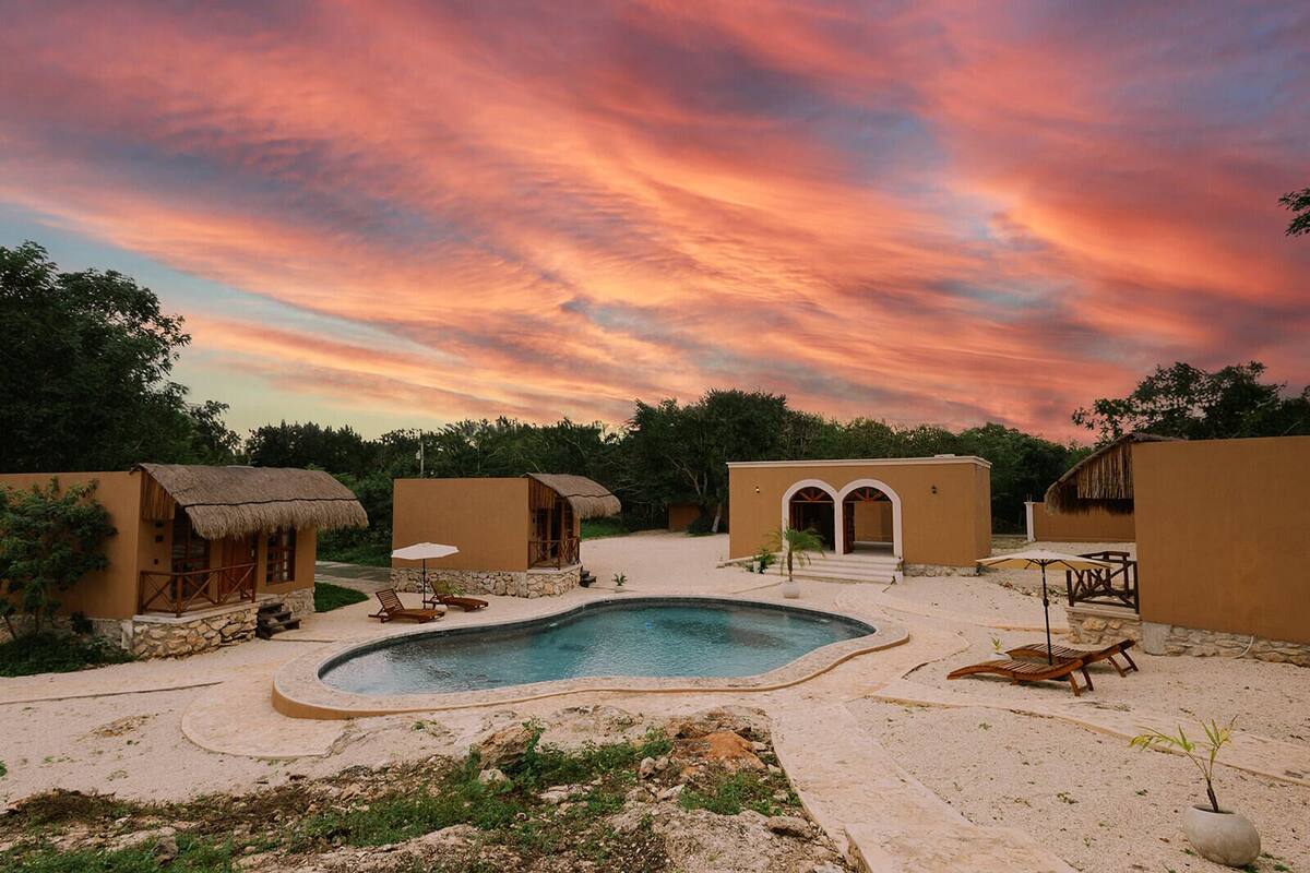 A resort with a swimming pool, thatched-roof huts, and a sunset sky.