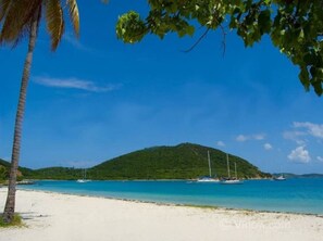 A beach with white sand, clear blue water, and a green hill in the background.