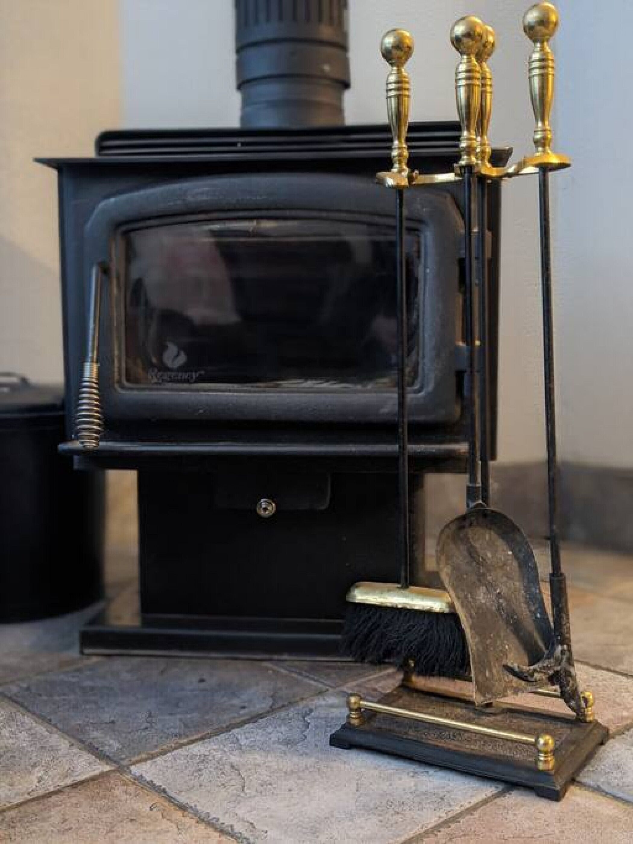 Ein schwarzer Kaminofen mit Schornstein, eine Metallbürste und eine Schaufel auf einem Fliesenboden.