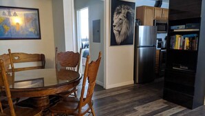 A dining area with a round wooden table and chairs, a kitchen with a refrigerator and cabinets, and a hallway leading to another room.