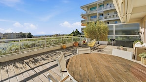 A wooden deck with a round table and chairs, potted plants, and a view of a building with balconies. - Appartement Margot - Welkeys (Le Cannet)
