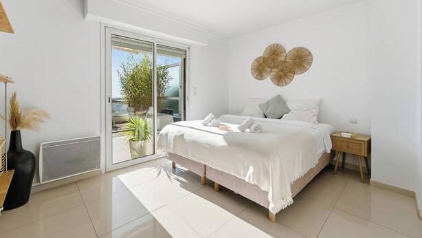 A bedroom with a bed, a bedside table, a radiator, a sliding glass door, and a view of the outdoors. - Appartement Margot - Welkeys (Le Cannet)