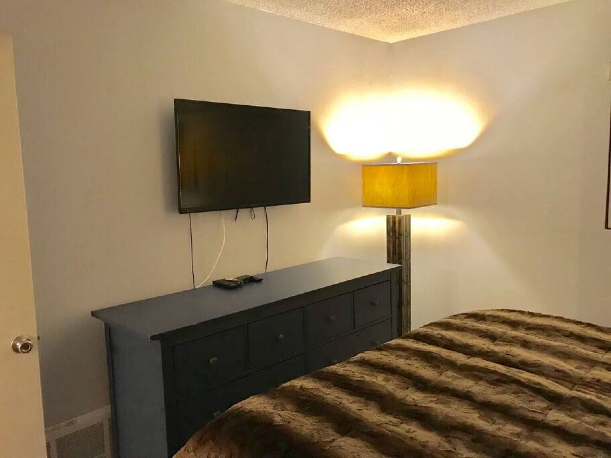 A bedroom with a bed, a dresser, a television mounted on the wall, and a bedside lamp.