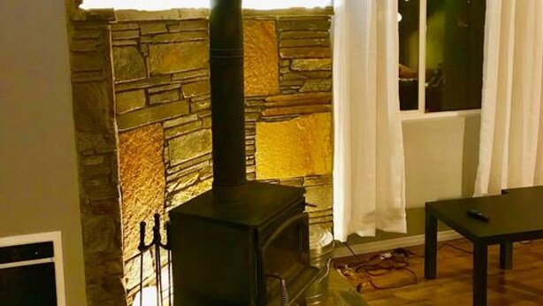 Une pièce chaleureuse avec une cheminée en pierre, un poêle en bois noir et une fenêtre ornée de rideaux blancs.