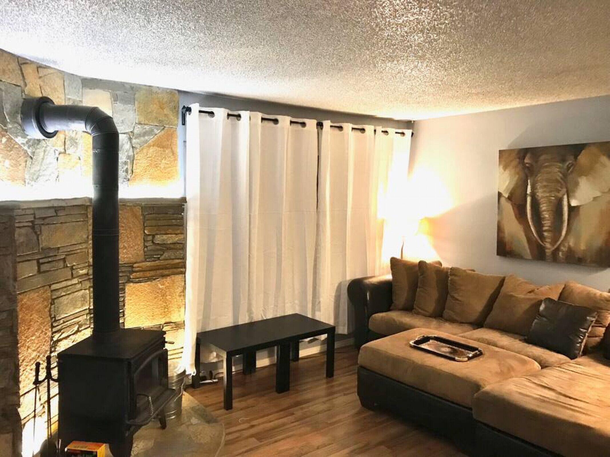 Ein gemütliches Wohnzimmer mit Kamin aus Stein, einem schwarzen Kaminofen, einem beigen Sofa, einem schwarzen Couchtisch und einem großen Elefantenbild an der Wand.
