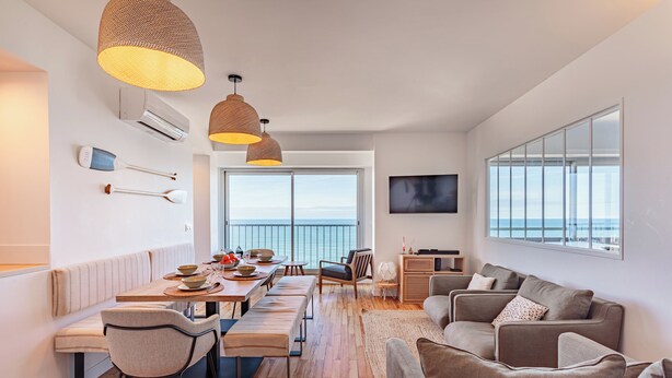 A modern living room with a large window, a dining area with wooden flooring, and a sofa set. - Appartement Okéana - 3 étoiles - Welkeys (Biarritz)