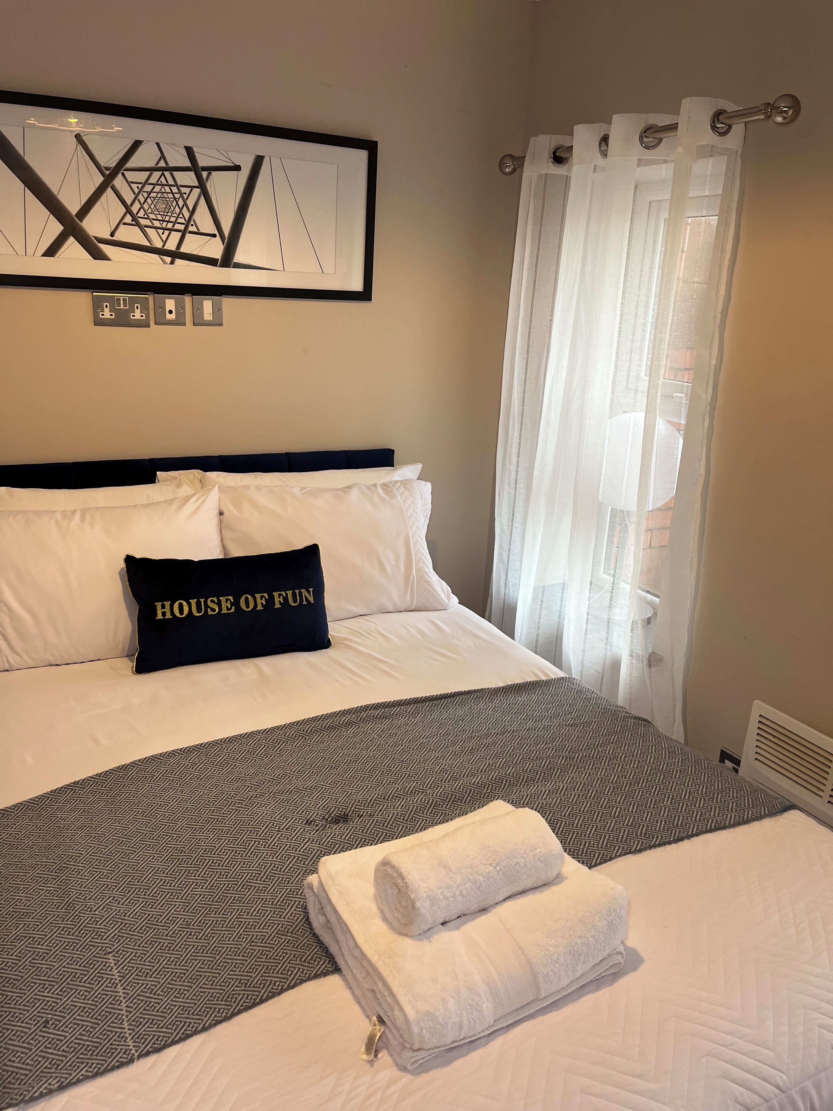 A neatly made bed with white bedding, a black pillow with "HOUSE OF FUN" text, a grey patterned blanket, and a folded white towel.