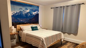 A bedroom with a bed, a nightstand, a lamp, a window with curtains, a wooden chair, and a wall-mounted radiator. - Cozy 3-bedroom house in Anchorage (Anchorage)