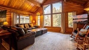 A log cabin interior with a wooden ceiling, large windows, a leather sofa, a television, and a fireplace. - The Log Cabin Where Memories are Made (Sunriver)