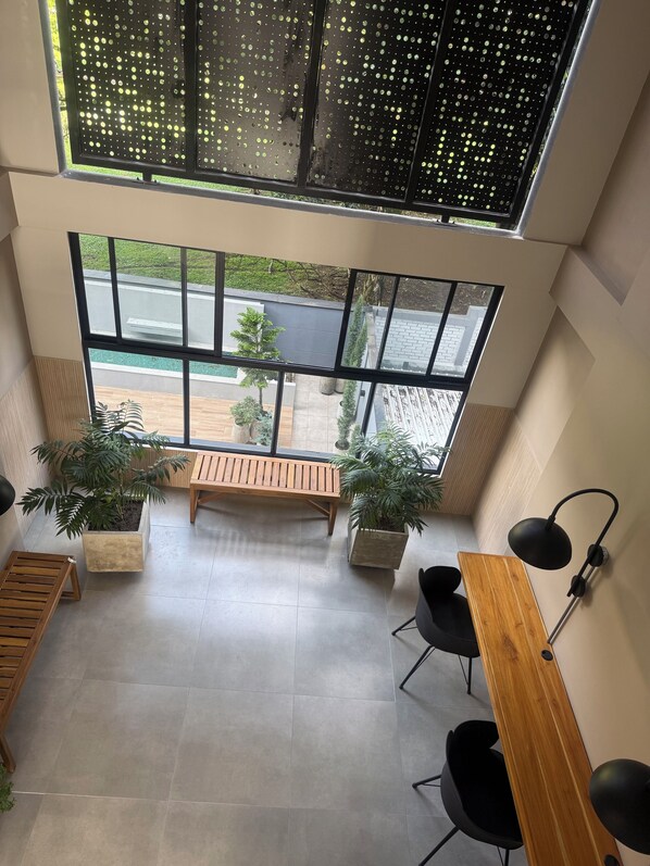 Un interior moderno con ventanas amplias, un banco de madera, plantas en macetas y una lámpara de escritorio negra.