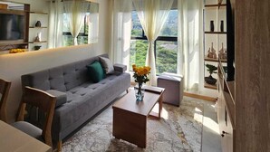 A modern living room with a grey sofa, wooden coffee table, and a view of greenery through large windows. - Cozy 1-bedroom apartment with AC in tranquil Cali (Cali)