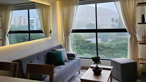 A modern living room with a sofa, coffee table, and dining table. Large windows offer a view of the cityscape and greenery. - Cozy 1-bedroom apartment with AC in tranquil Cali (Cali)