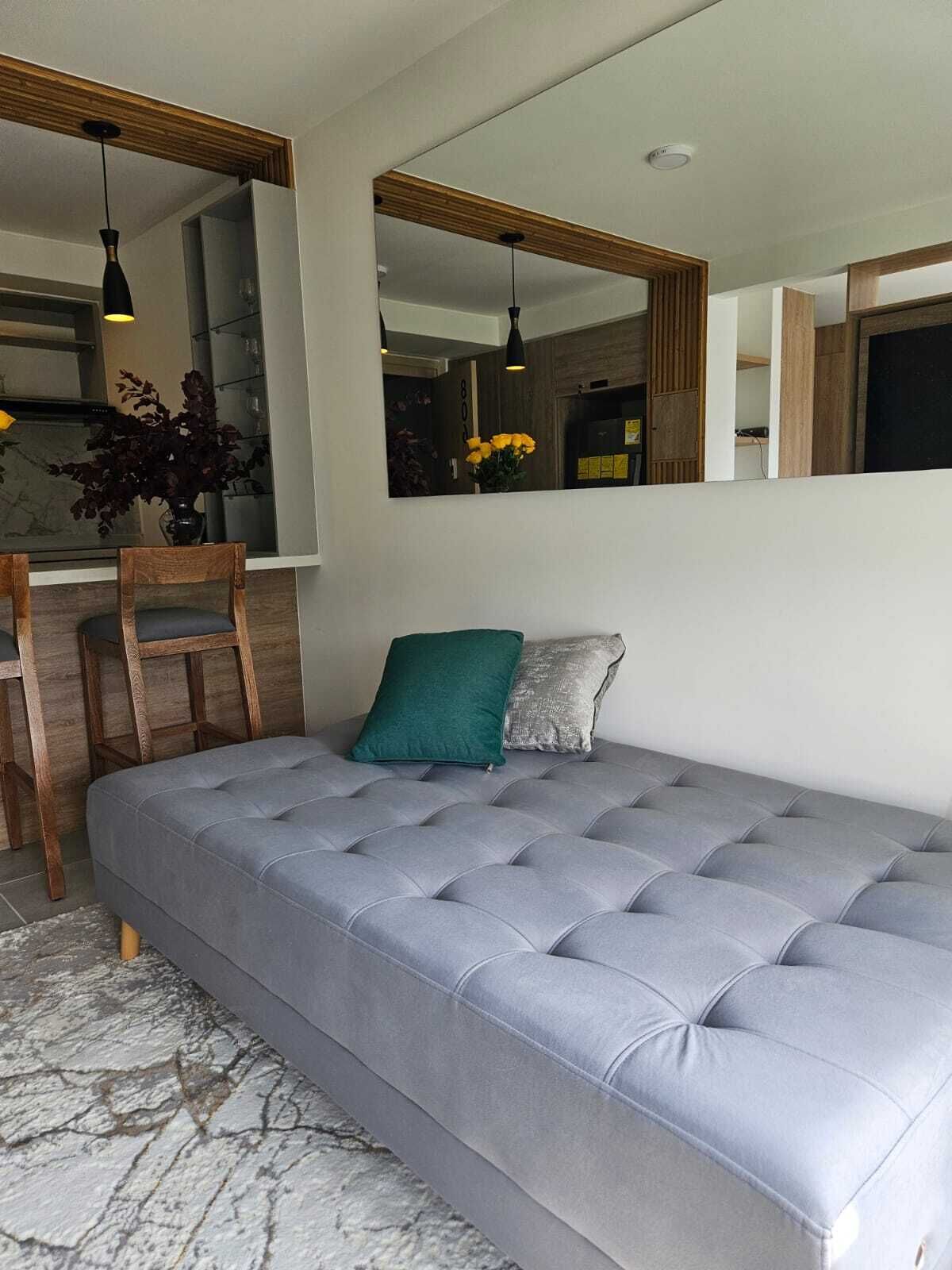 Una sala moderna con un sofá gris, una silla de madera y una mesita. Hay dos espejos en la pared, y se ve una planta al fondo.