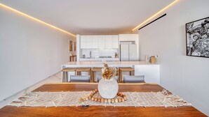 A modern kitchen with white cabinetry, a central island, and a dining area with a wooden table and woven placemat. - Beautiful New Luxury Apartment - Cora Sunset at Campestre (San Jose Del Cabo)