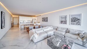 A modern living room with a large grey sectional sofa, a marble coffee table, and a kitchen in the background. - Beautiful New Luxury Apartment - Cora Sunset at Campestre (San Jose Del Cabo)