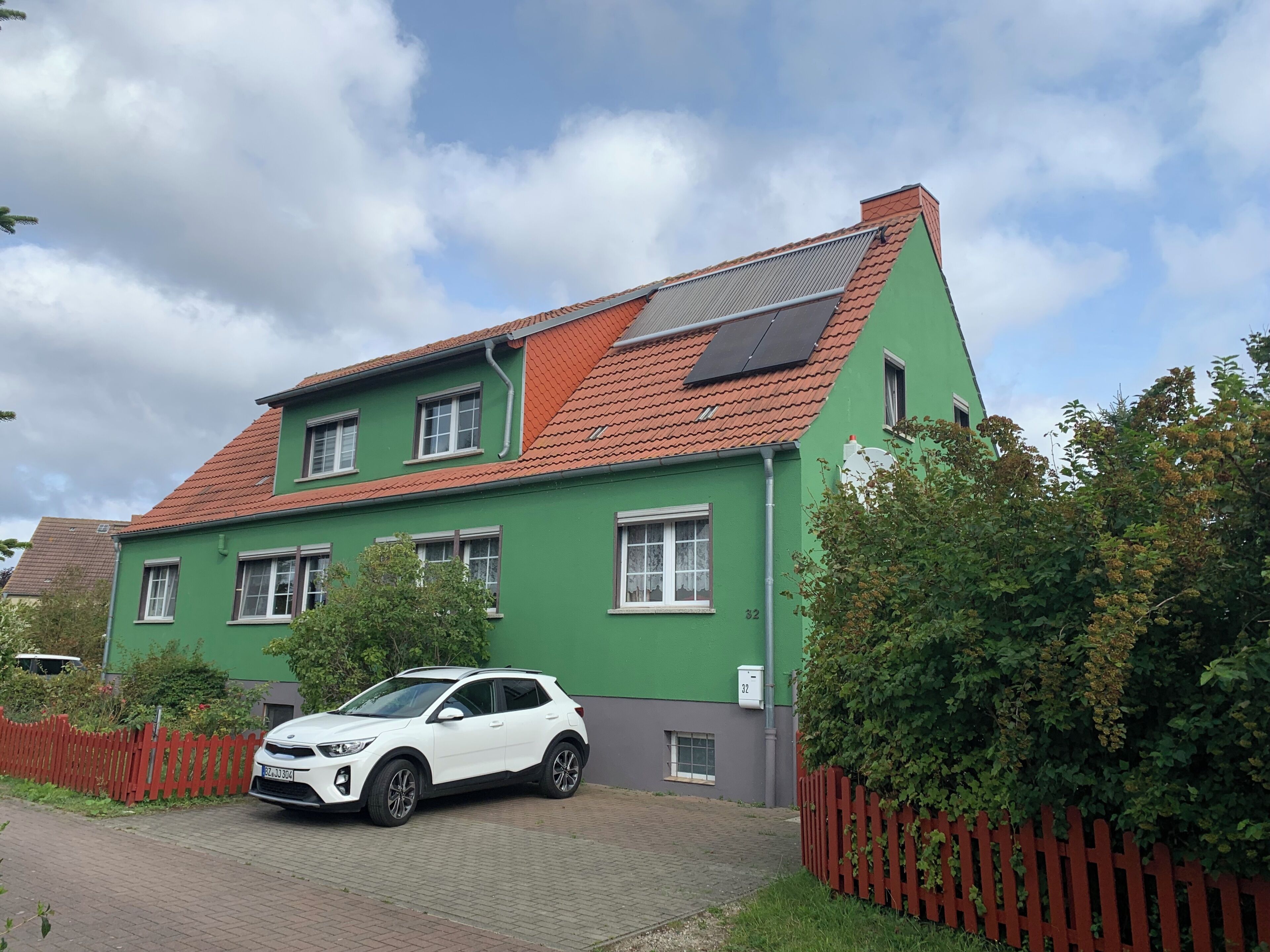 A green house with a red roof, solar panels, and a white car parked in front.