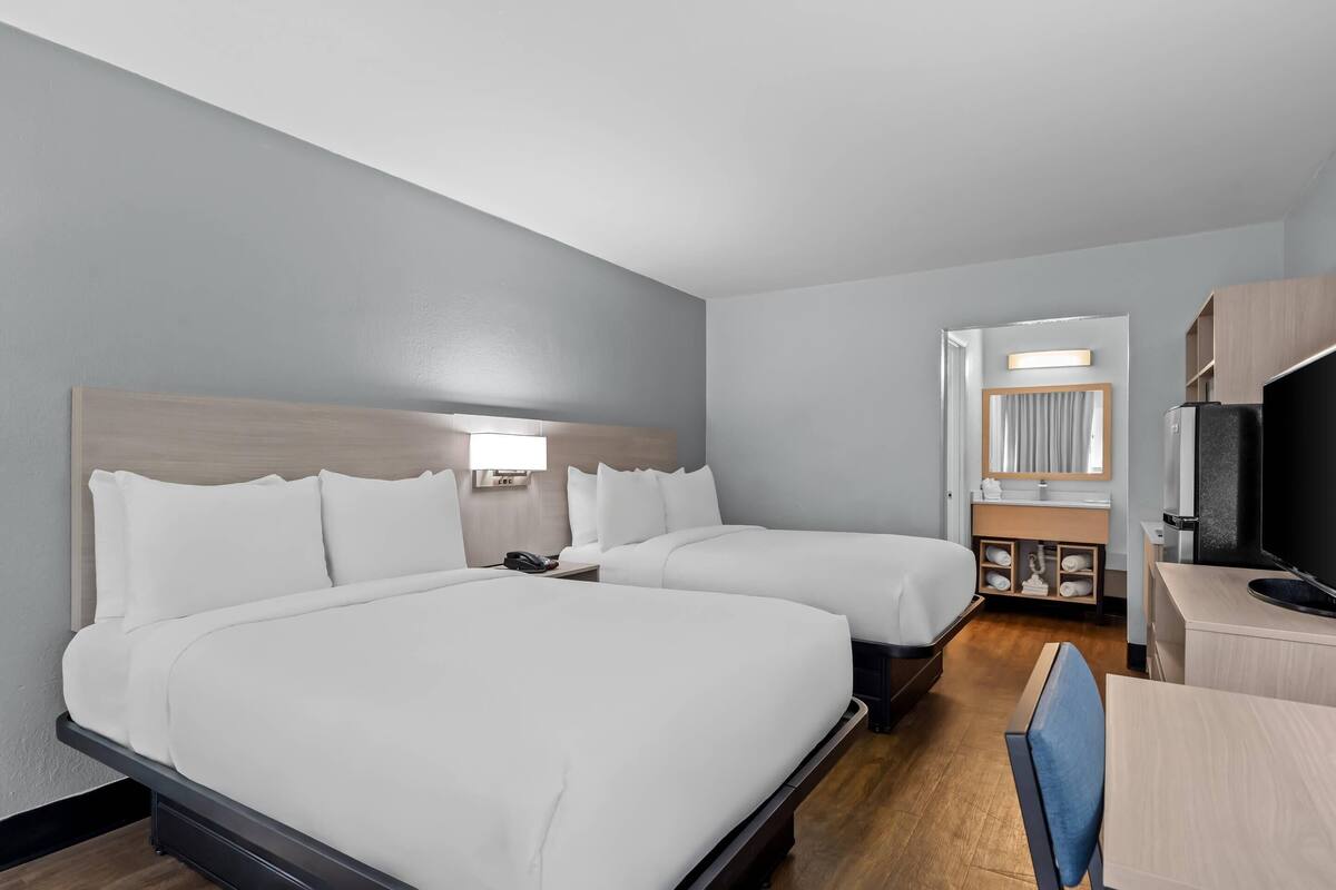 Ein Hotelzimmer mit zwei Betten, einem Schreibtisch, einem Fernseher und einem durch eine offene Tür einsehbaren Badezimmer.